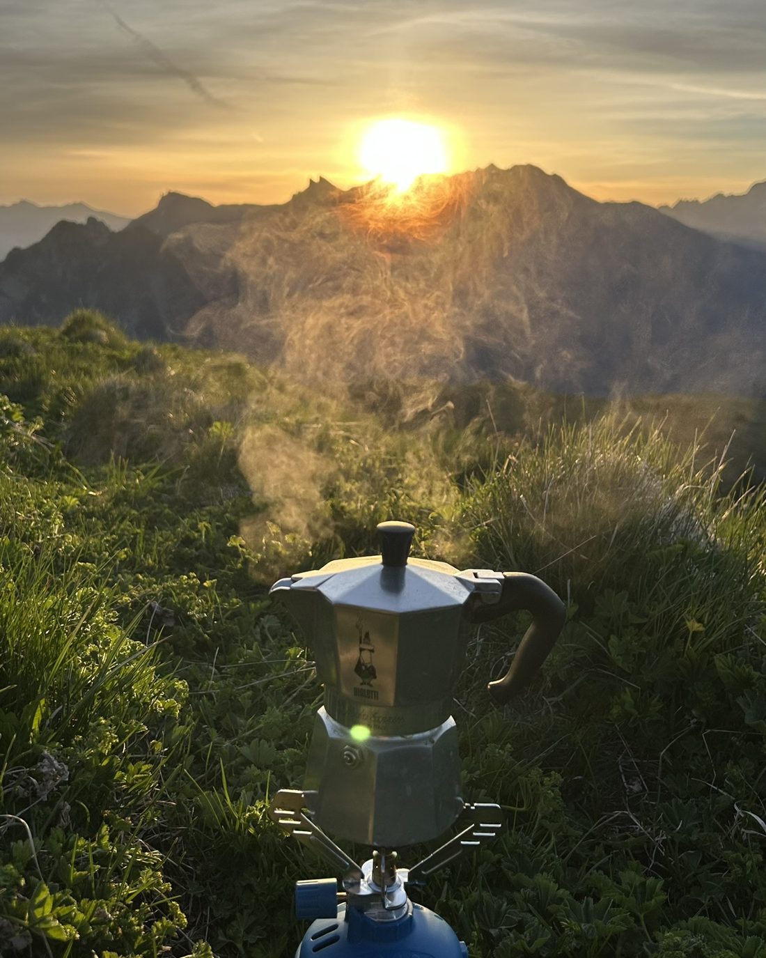 Dampfende Bialetti auf einem Gaskocher, dahinter geht die Sonne über einem Berggrat auf